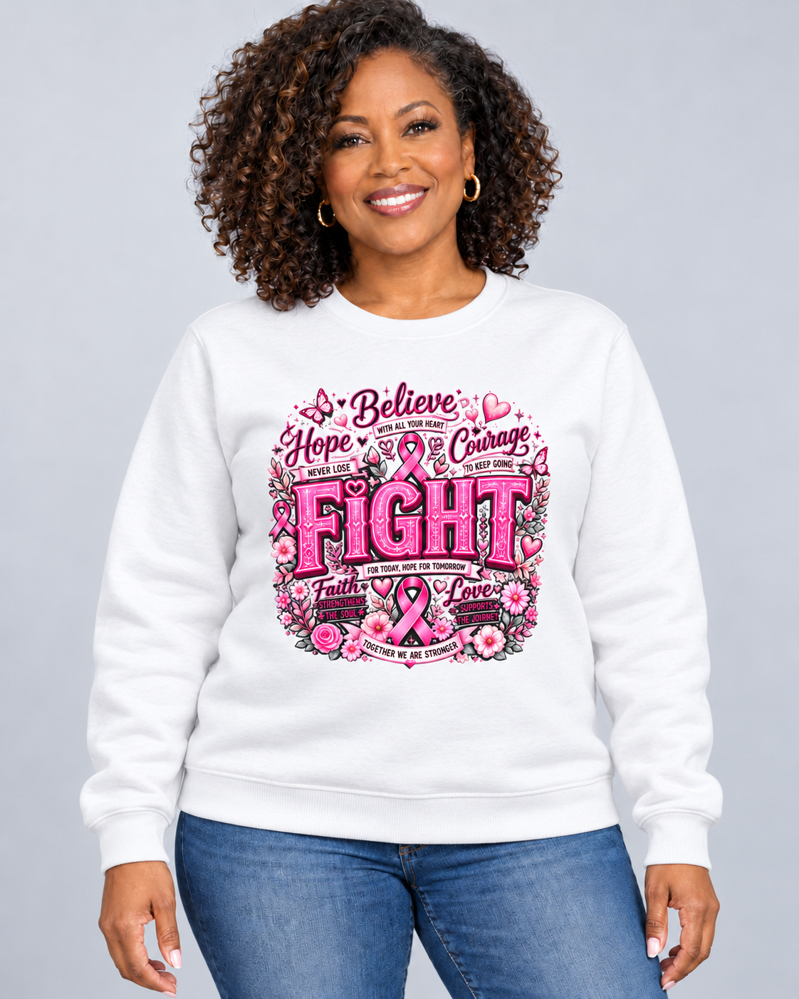A smiling woman with curly hair wearing a white crewneck sweatshirt with a pink and black breast cancer awareness graphic that reads 'Believe Hope & Courage FIGHT faith love' surrounded by flowers and ribbons, paired with blue jeans against a light gray background.