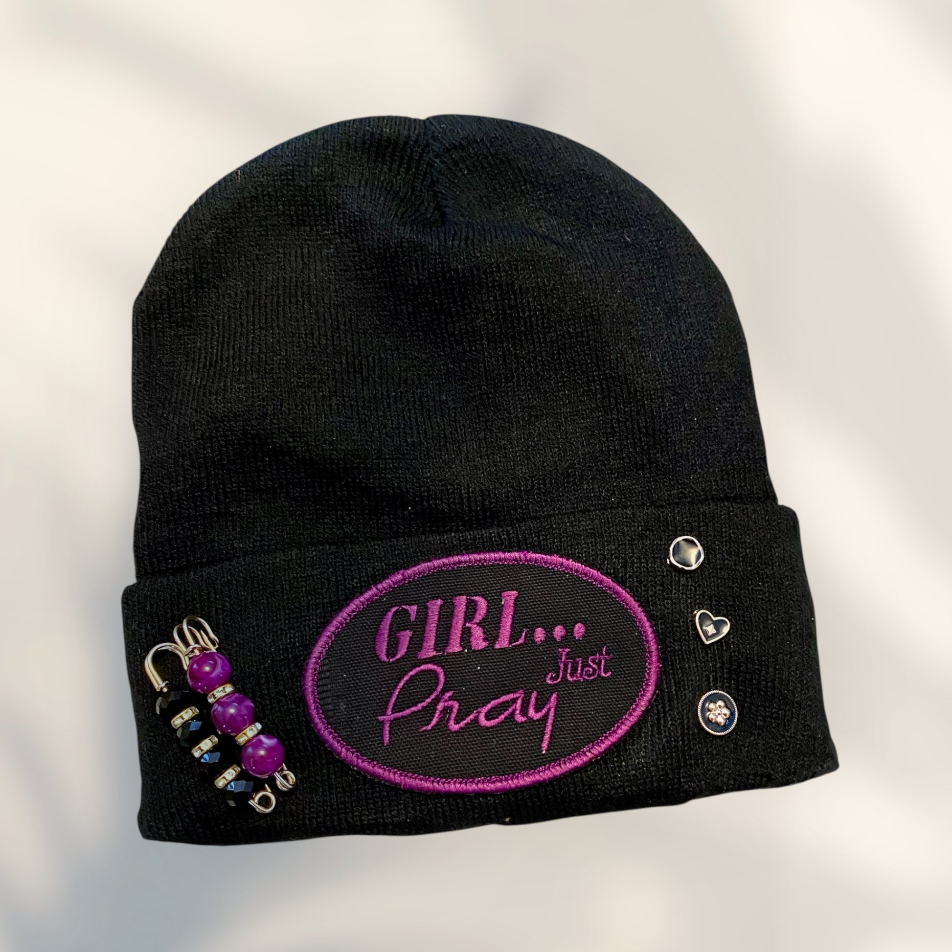 Black beanie with pink 'Girl Just Pray' text and decorative elements on a light gray background