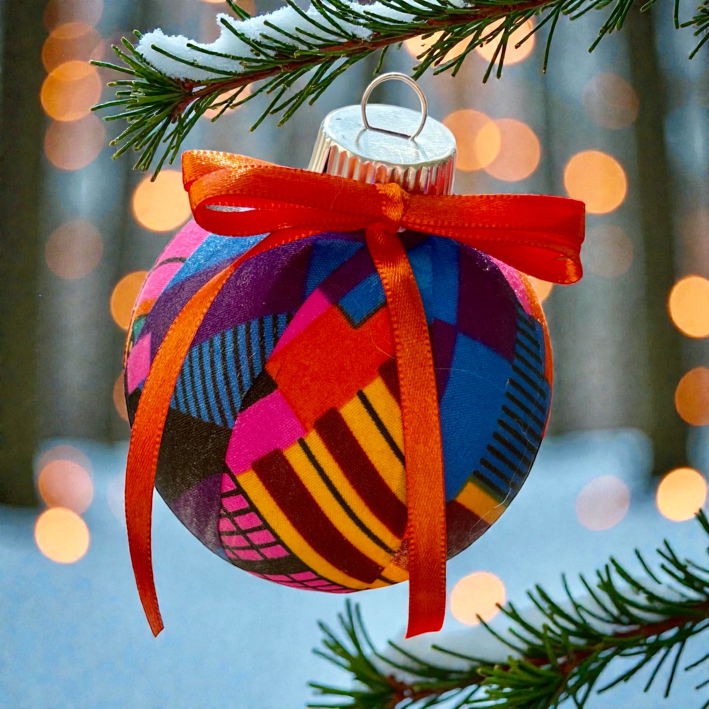 Colorful Christmas ornament with a red ribbon on a tree branch with blurred lights in the background